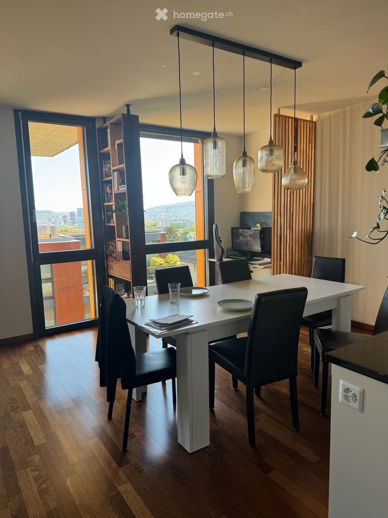 Modern dining area with a white dining table, six black chairs, pendant lights, and a laptop on a nearby desk. Large windows offer a scenic view.