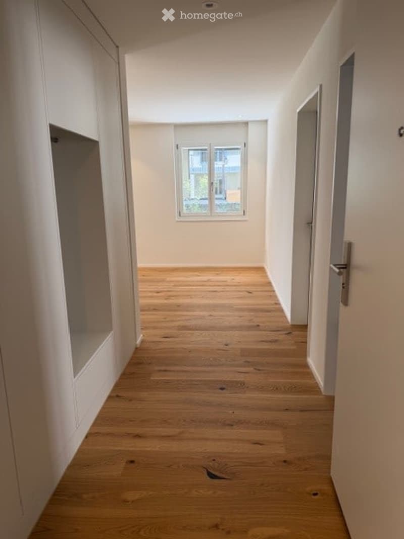 Empty hallway with light wooden flooring, white walls, and a single window at the far end.