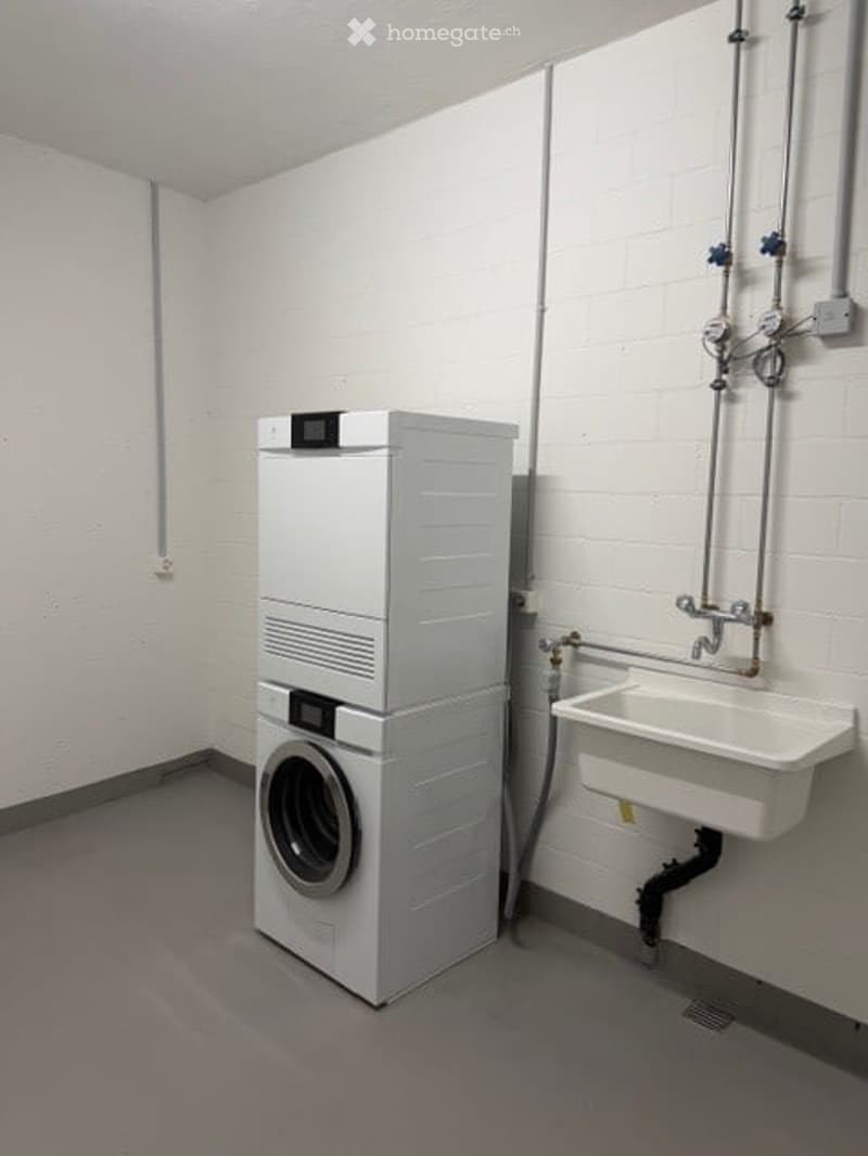 A compact bathroom with a stacked washing machine and a small sink, white tiled walls, and a grey floor.