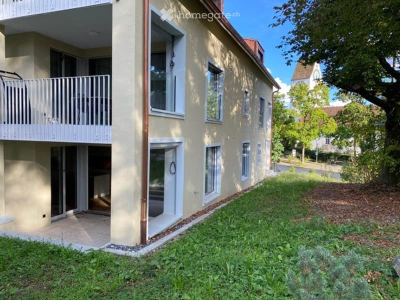 Two-story residential building with a balcony on the upper floor, surrounded by greenery and a paved pathway.