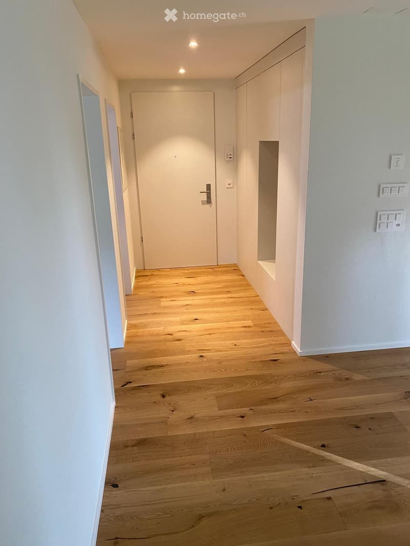 Empty hallway with light wooden flooring, white walls, and a closed door at the end.