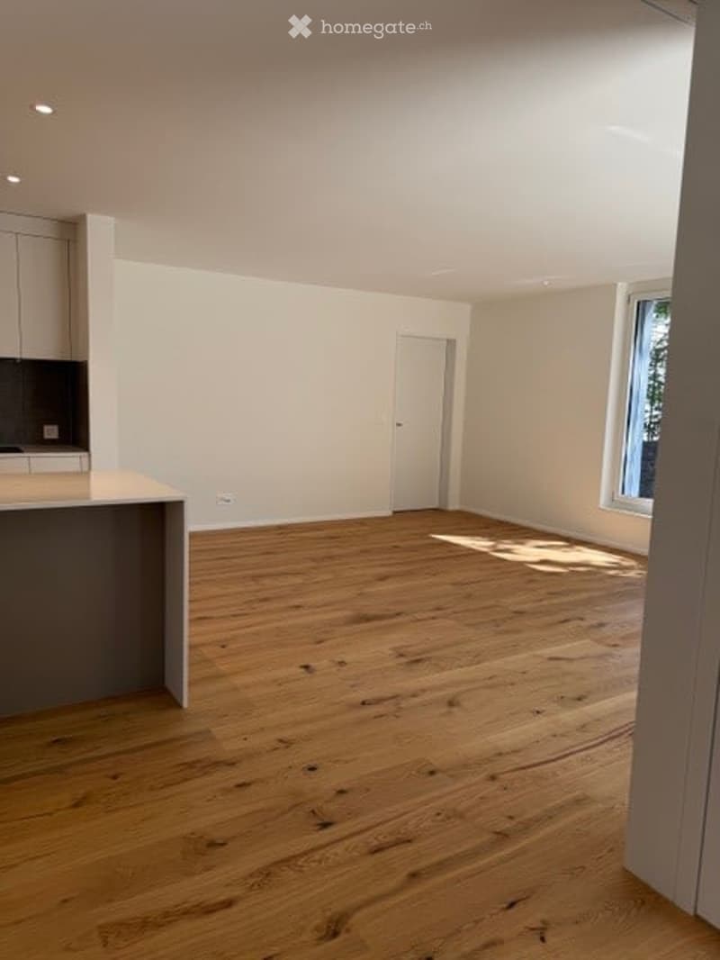 Empty room with wooden flooring, white walls, and a small kitchen area visible in the background.