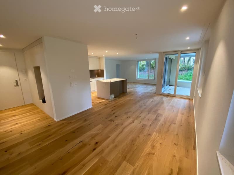 Empty room with light wooden flooring, white walls, and recessed lighting. Features a sliding glass door leading to an outdoor area and a small kitchen area visible in the background.