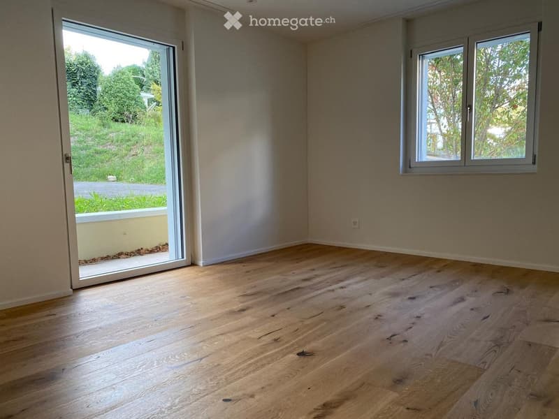 Empty room with light wooden flooring, white walls, large sliding glass door leading to a green outdoor area, and a window with white frame.