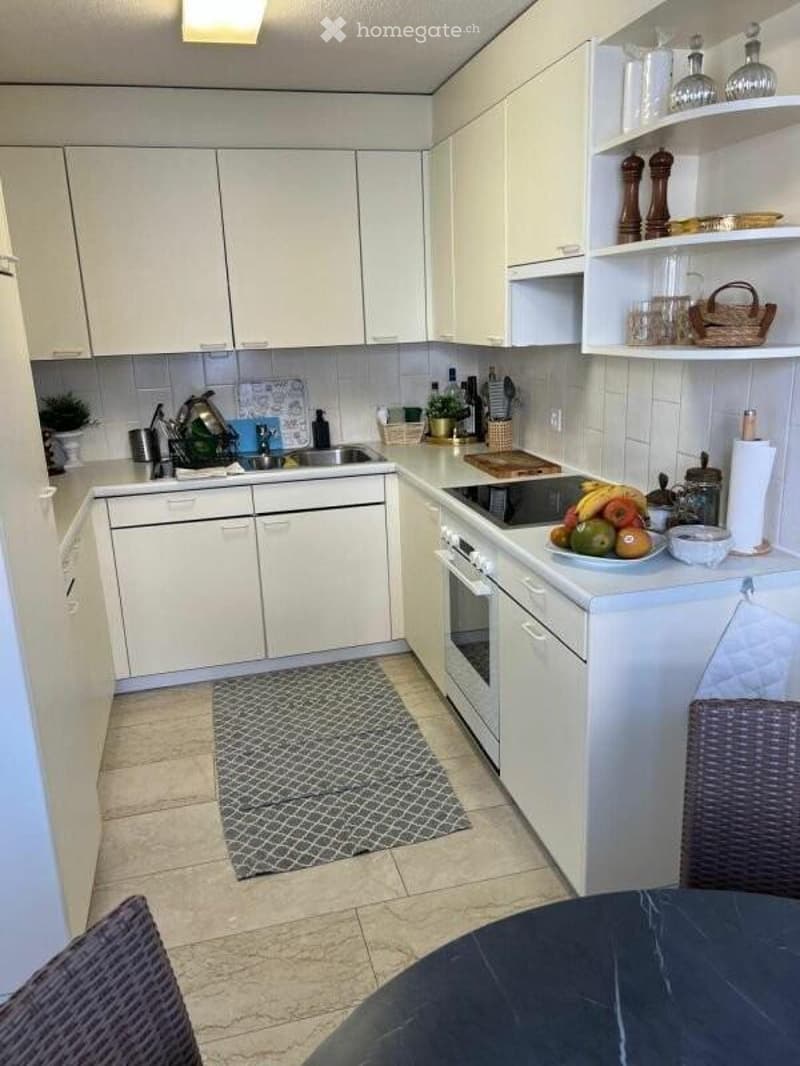 Modern kitchen with white cabinets, stainless steel appliances, and a tiled floor. The counter features a bowl of fruit, a cutting board, and various kitchen items.
