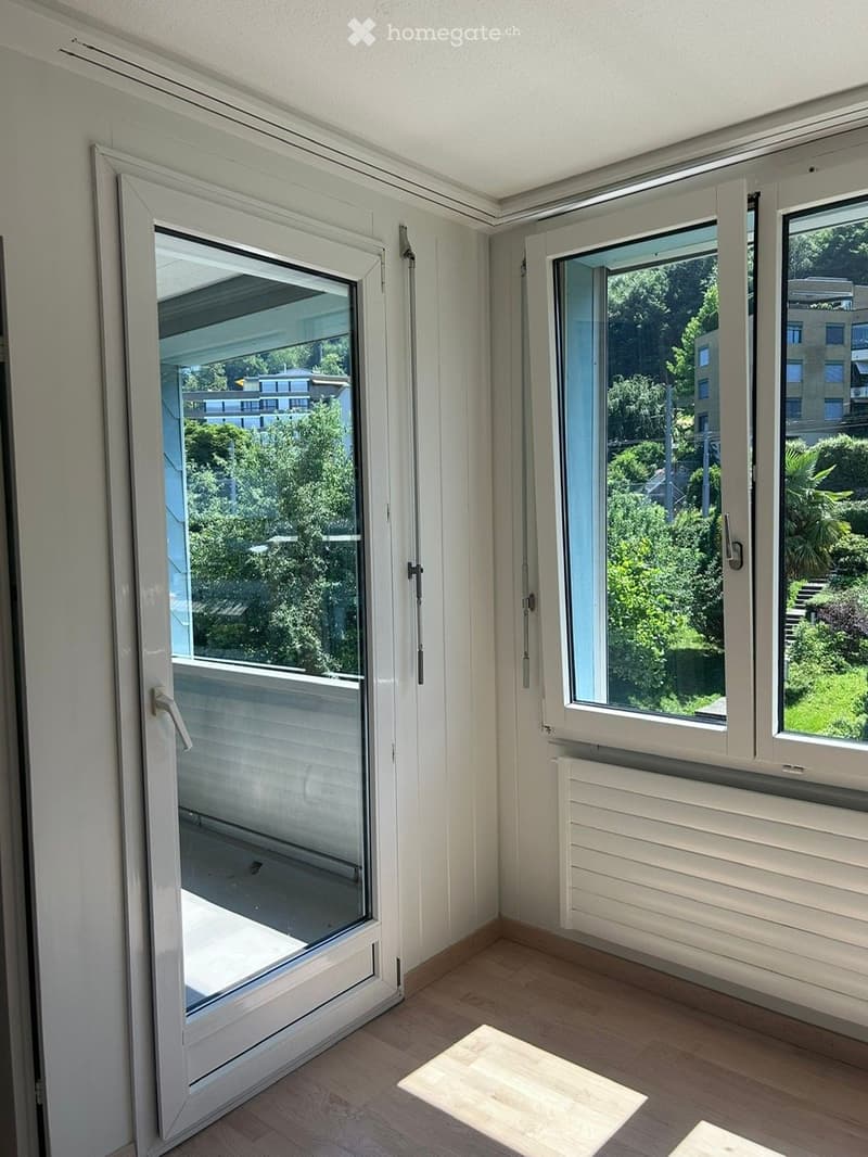 Empty room with wooden flooring, large windows, and a sliding glass door leading to a balcony. The room has white walls and a radiator under the windows.