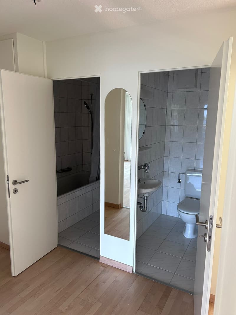 Bathroom with a toilet, sink, and bathtub, visible through an open door. The floor is wooden, and the walls are tiled.
