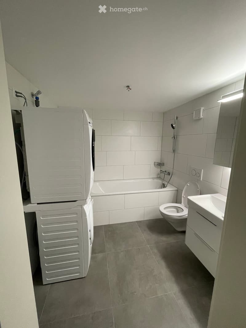 Modern bathroom with a bathtub, toilet, sink, and built-in cabinet. Light grey tiles and neutral tones.