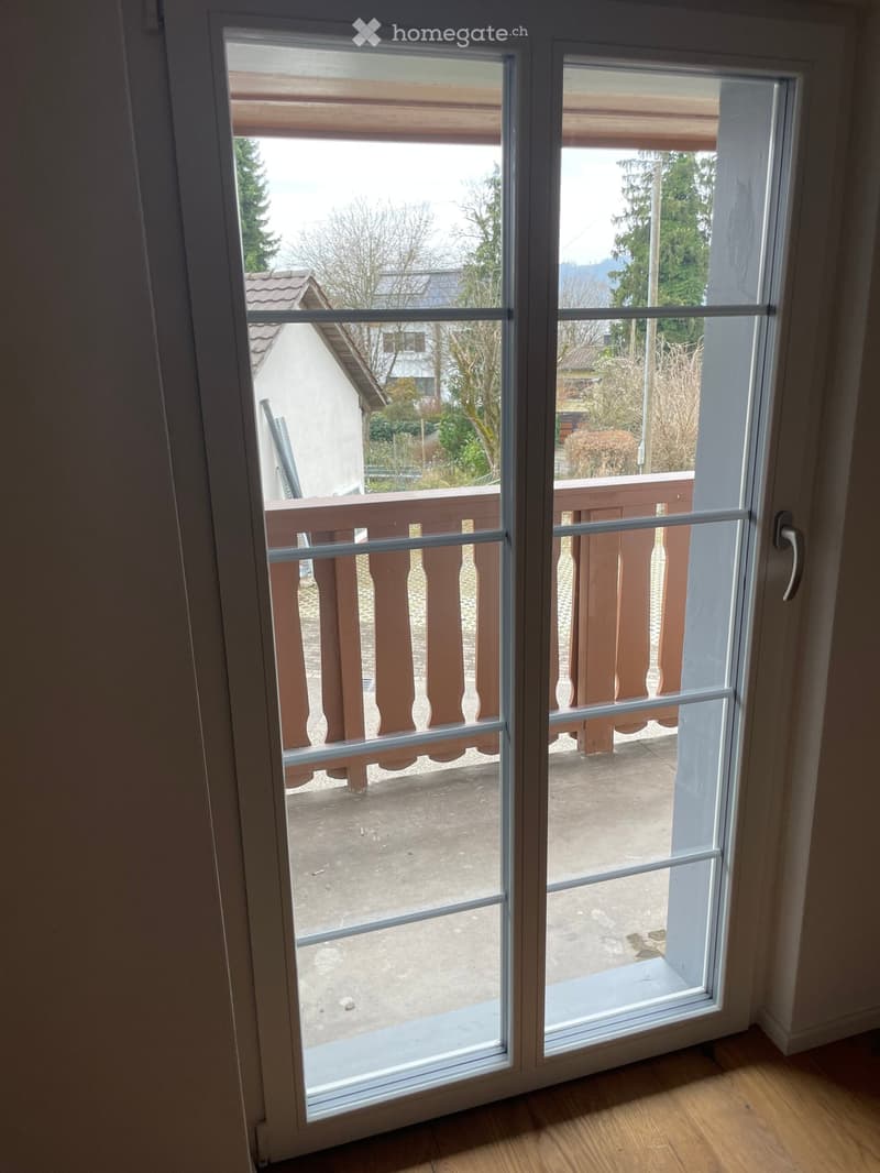 Sliding glass door opening to a wooden balcony with a railing, overlooking a residential area with trees and houses.