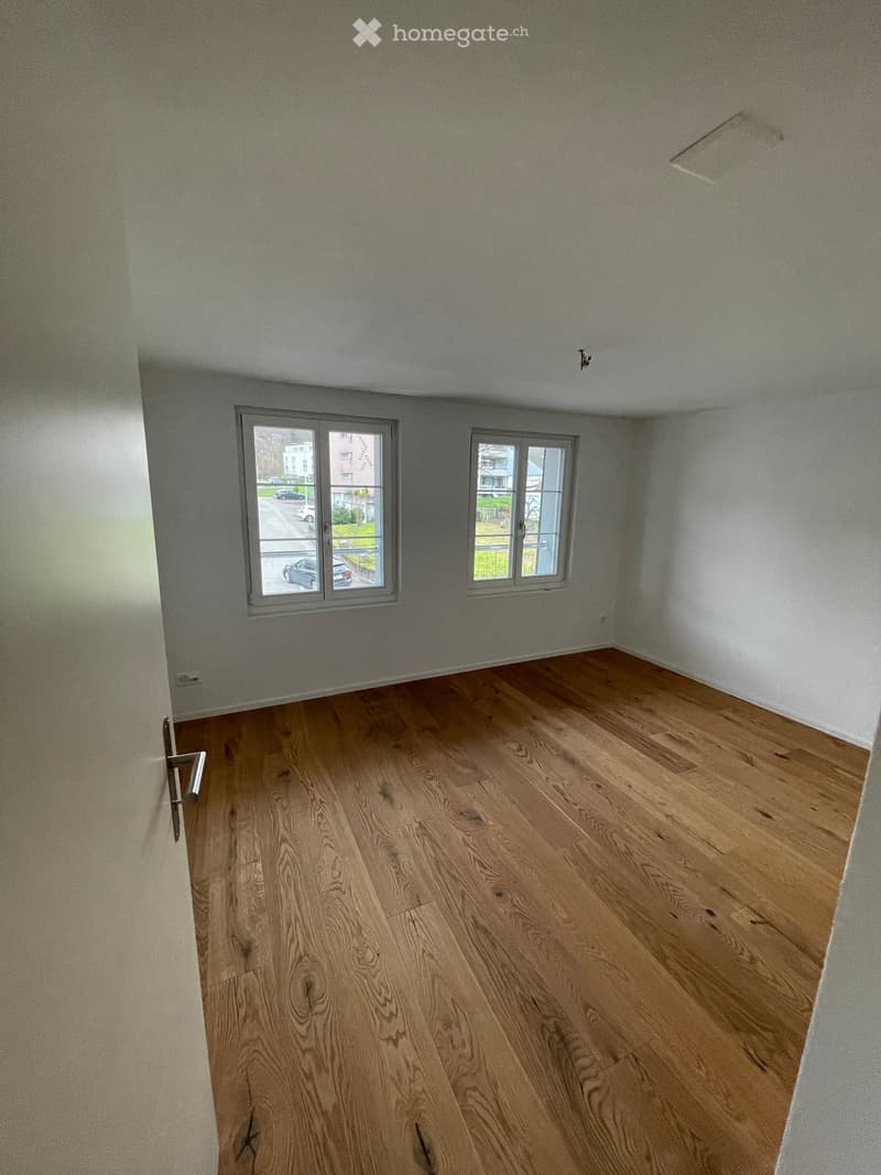 Empty room with wooden flooring and white walls, featuring two windows and a door.