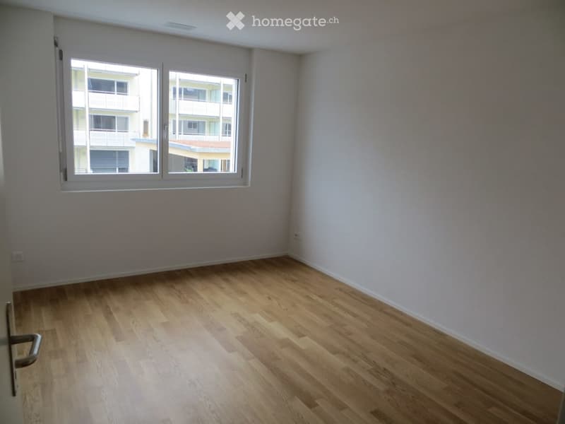 Empty room with wooden flooring and large windows, showing natural light and a view of neighboring buildings.