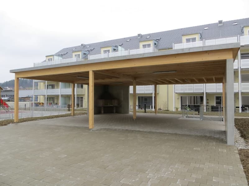 Modern multi-story building with a covered parking area featuring wooden beams and concrete pillars.