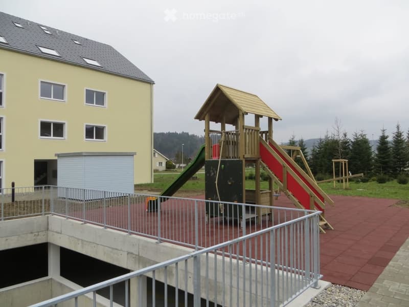 Outdoor playground area with slides, climbing structures, and a small wooden playhouse, surrounded by a metal railing and adjacent to a yellow building.