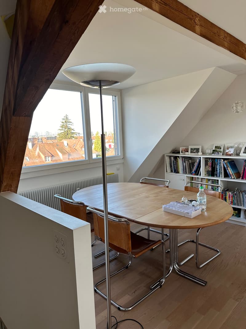 Bright attic room with wooden floors, large windows offering a city view, round wooden table with chairs, bookshelf, and floor lamp.
