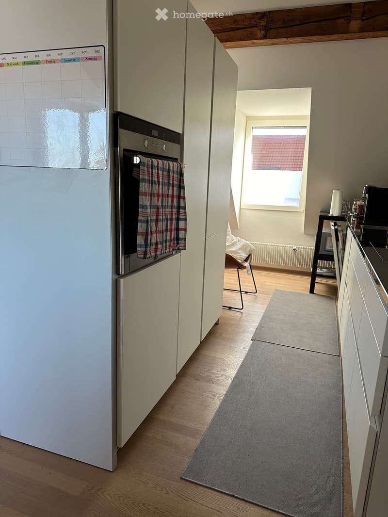 A modern kitchen with a refrigerator, oven, and a small living area with a chair and a stool. The floor is wooden, and there is a grey rug in the kitchen area.