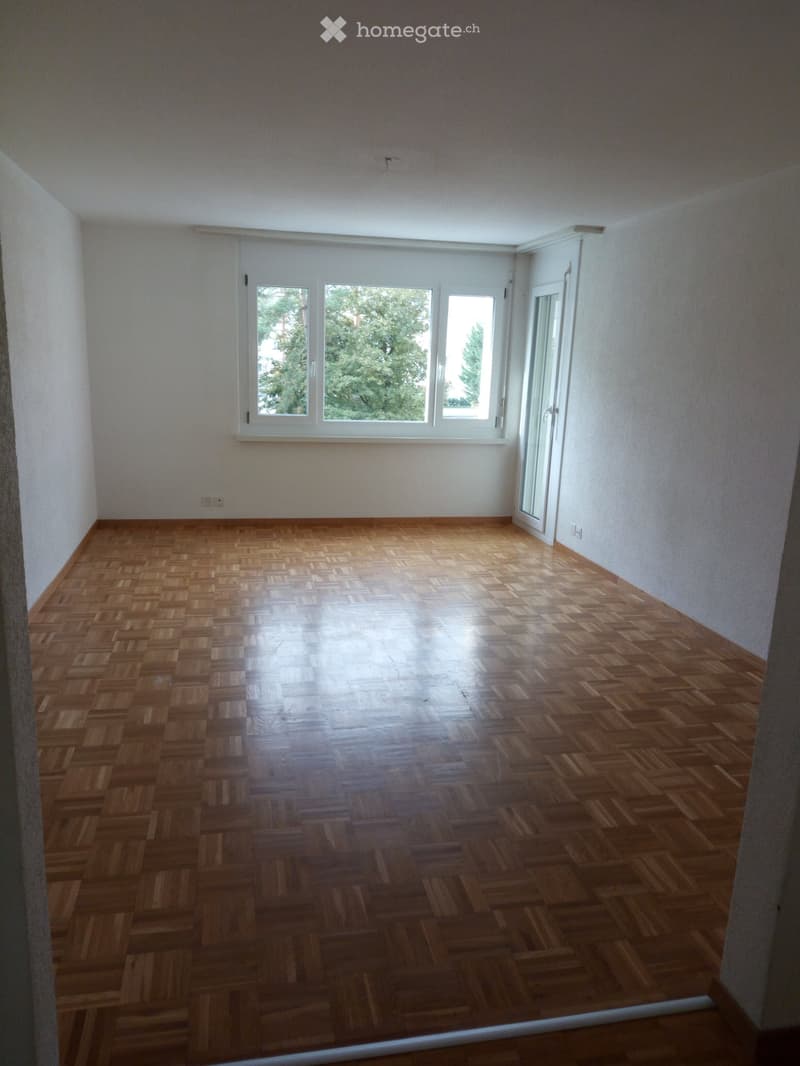 Empty room with wooden parquet flooring, white walls, large windows with white frames, and a sliding glass door leading outside.