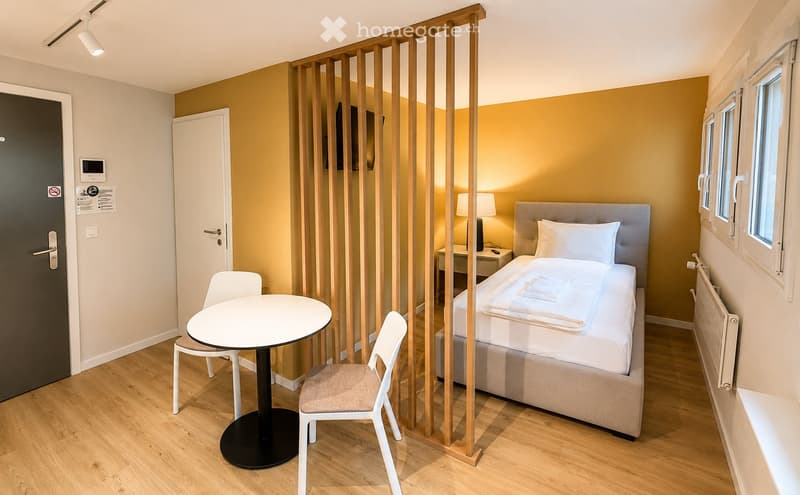 Cozy, well-lit bedroom with a bed, a small table, chairs, and a lamp. The room features a wooden floor, warm yellow walls, and a modern wooden partition separating the sleeping area from the seating area.