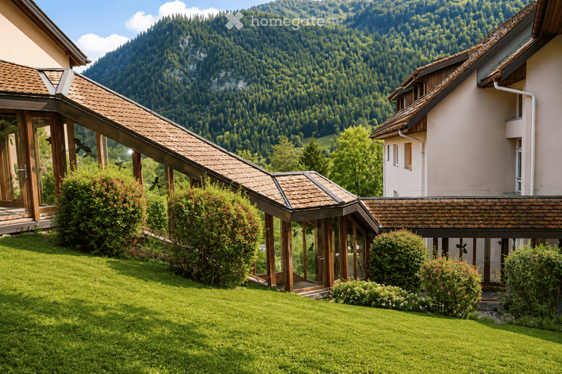 A two-story house with a tiled roof, surrounded by a well-maintained lawn and lush greenery, with a scenic mountain backdrop.