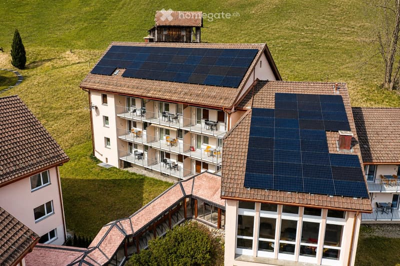 Aerial view of a multi-story residential building with solar panels on the roof, surrounded by green fields and trees. Balconies with outdoor furniture are visible on the upper floors.
