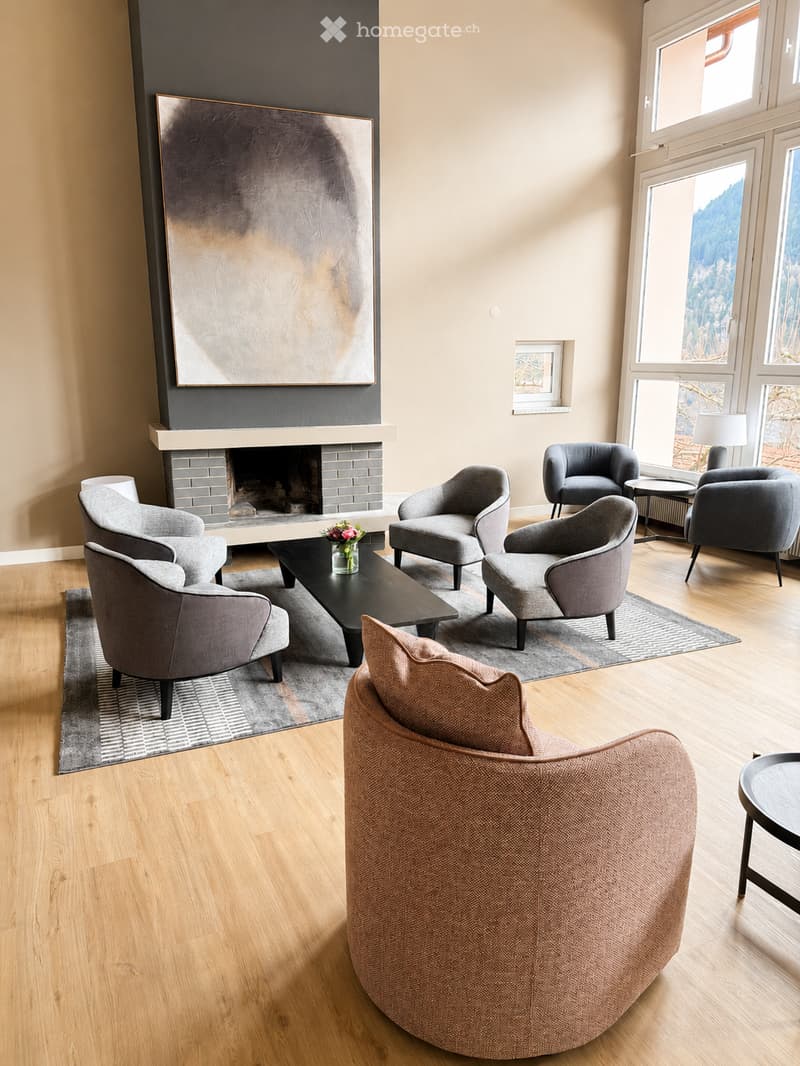 Modern living room with large windows offering a scenic view, a fireplace, grey armchairs, a sofa, coffee table, and a side table with a lamp.