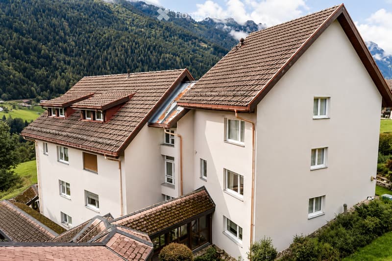 A multi-story white building with a tiled roof, multiple windows, and a scenic mountain backdrop.