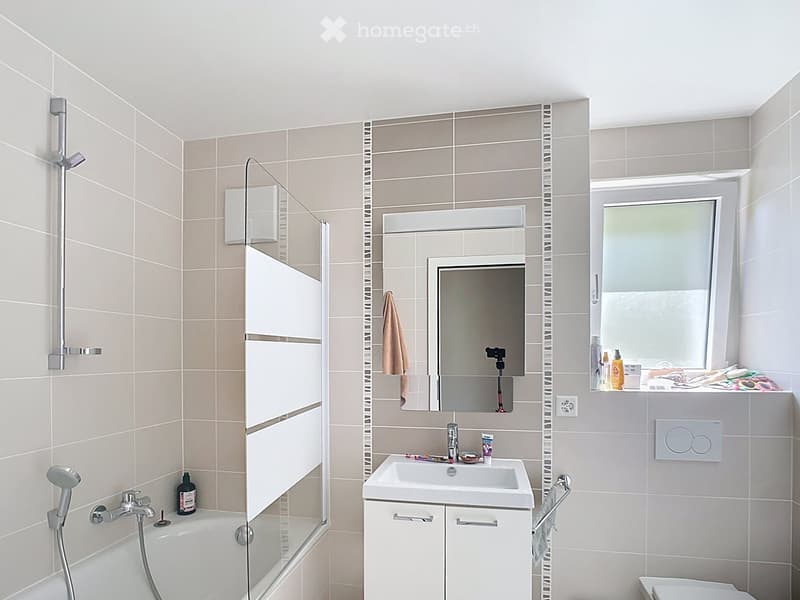 Modern bathroom with a bathtub, shower, sink, mirror, and cabinet. The walls are tiled in beige and grey, and there is a window with frosted glass.