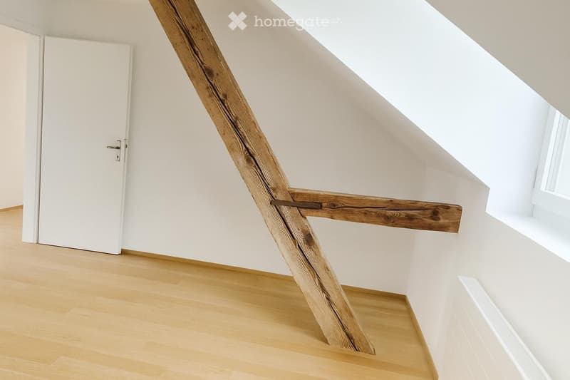 Modern hallway with wooden floor, white walls, and a unique wooden staircase railing. Features a white door and a window letting in natural light.