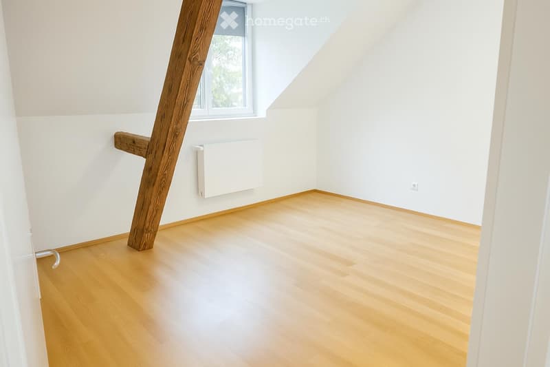 Empty room with light wooden flooring, white walls, and a large window with a view of greenery. A wooden structural beam is visible, and a radiator is mounted on the wall.