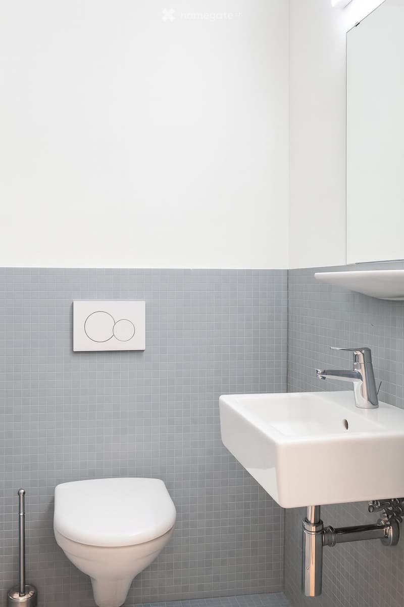 Modern bathroom with grey tiled walls, white toilet, white sink with chrome faucet, and a toilet brush holder.