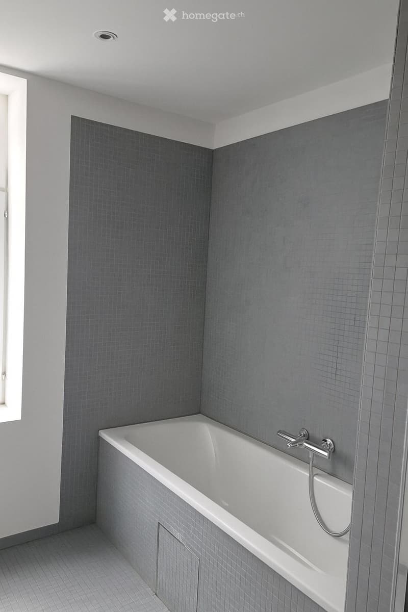 Modern bathroom with a white bathtub, grey tiled walls, and a built-in shower handle.