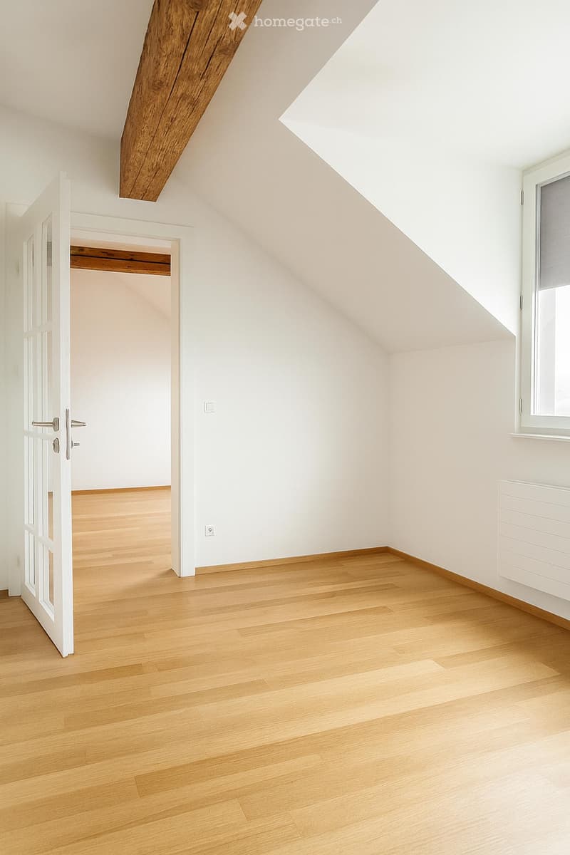 Empty attic room with wooden floor, white walls, exposed wooden beams, and a window with blinds. A white door leads to another room with a closet visible.
