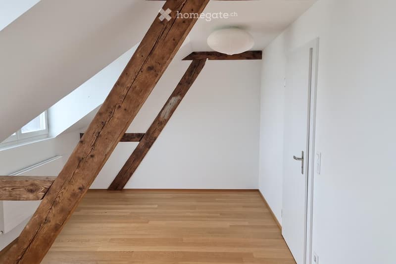 Empty attic space with wooden floor, white walls, exposed wooden beams, and a single light fixture on the ceiling.