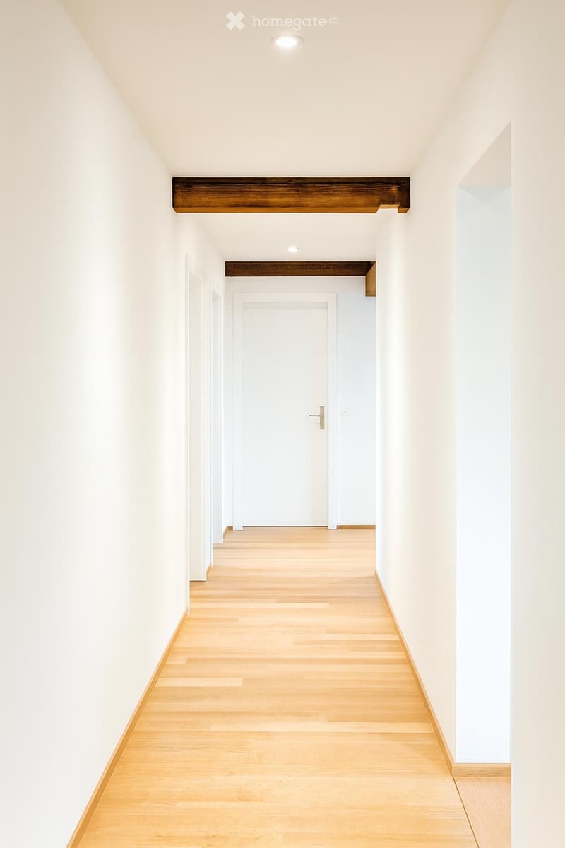 Empty hallway with light wooden flooring, white walls, and recessed lighting. Features a closed door at the end and exposed wooden beams on the walls.