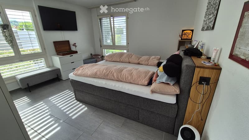 Bright bedroom with a large bed, nightstand, chest of drawers, and television mounted on the wall. Natural light streams in through large windows with blinds.