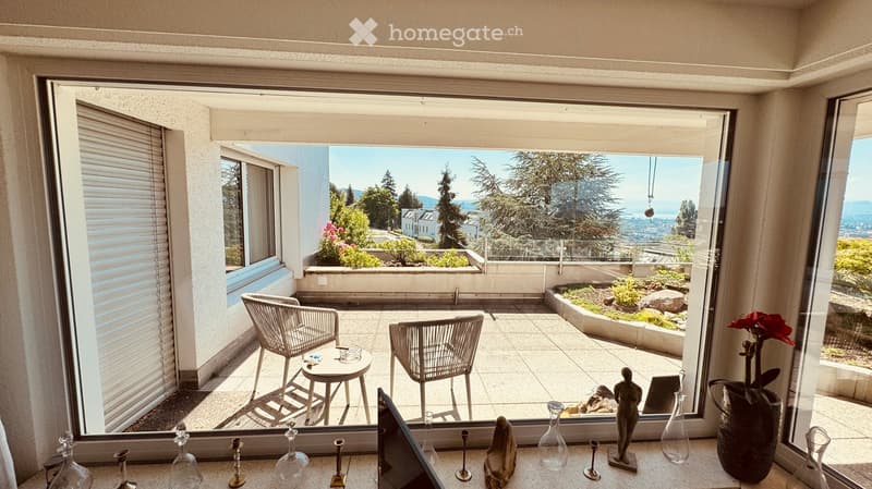 Balcony with a view of the city and mountains, featuring a small table and two chairs, surrounded by greenery and decorative items on the windowsill.