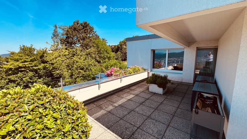 Balcony with tiled floor, glass railing, potted plants, and scenic view of trees and cityscape.