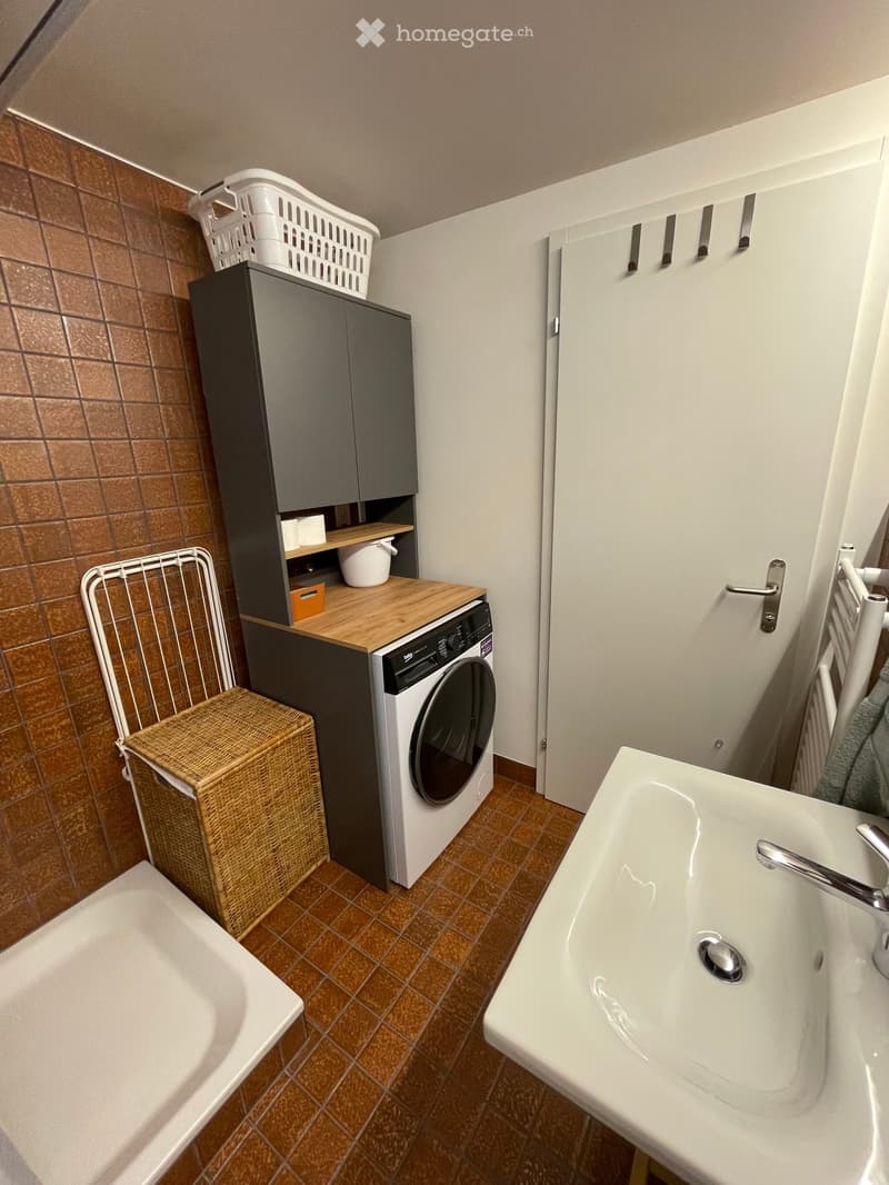 Bathroom with a sink, bathtub, washing machine, and a wicker basket. The walls are tiled, and there is a cabinet above the washing machine.