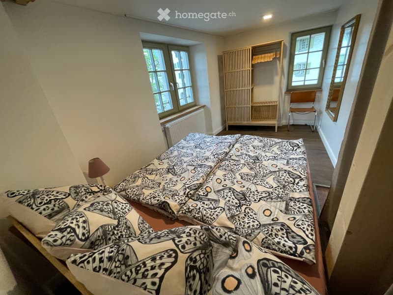 Cozy bedroom with a large bed covered in a patterned duvet, a bedside lamp, a wooden wardrobe, and a chair near the window.