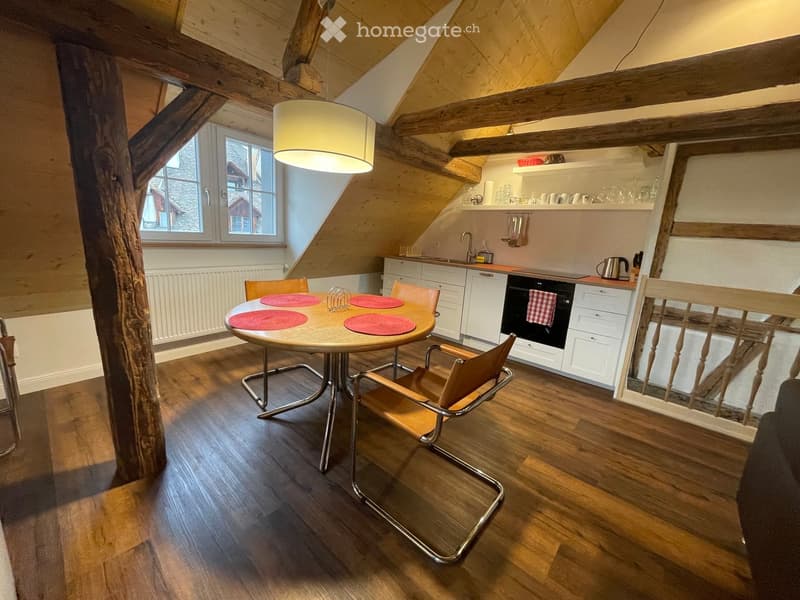 A cozy attic kitchen with wooden beams, a round wooden dining table with four chairs, white cabinets, a black oven, and a hanging pendant light.
