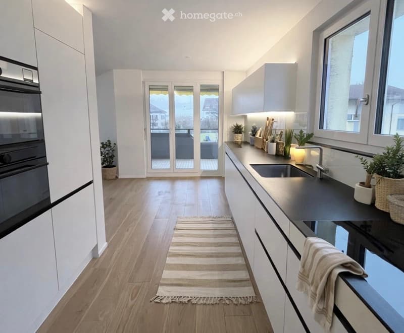 Modern kitchen with white cabinets, dark countertops, built-in oven, sink, and small plants on the counter. A sliding glass door leads to a balcony.