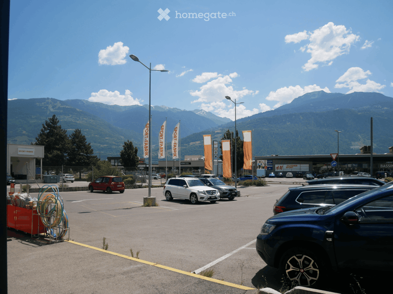 Parking lot with several cars, surrounded by trees and mountains, with commercial buildings and flags visible in the background.