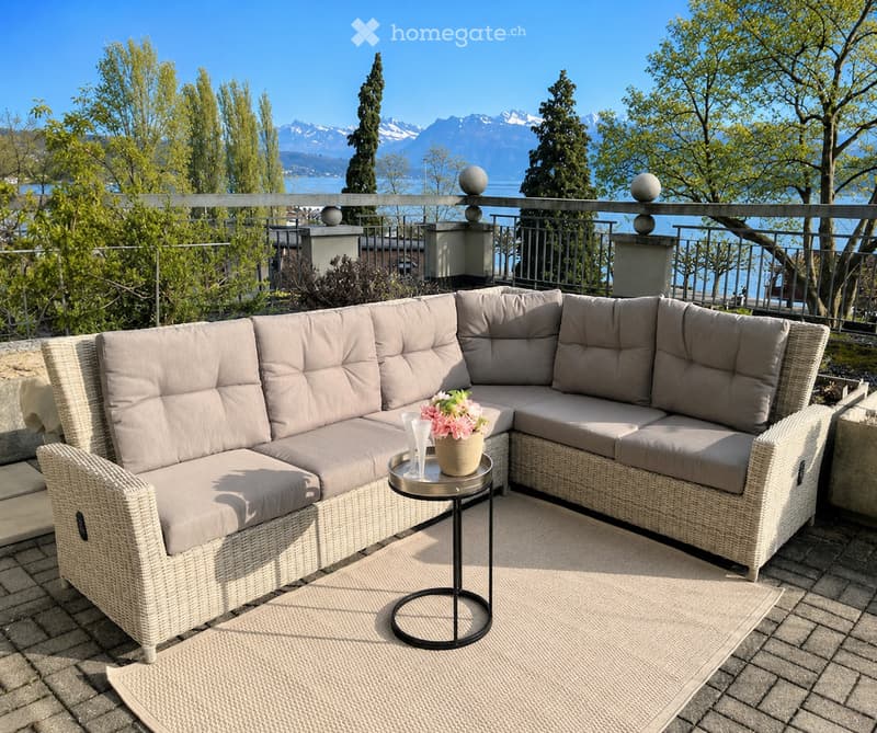 Balcony with a wicker sofa set, small round table, and flower pot, overlooking a scenic mountain and lake view.