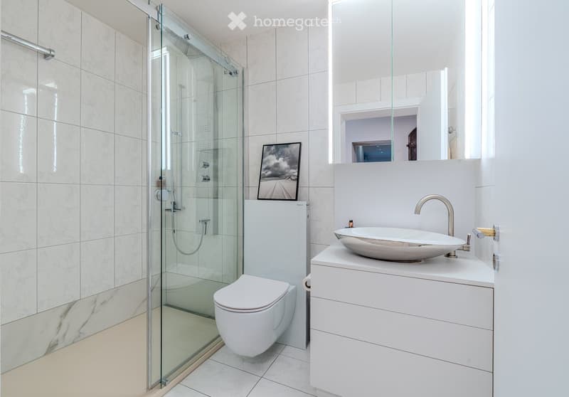Modern bathroom with white tiles, a glass-enclosed shower, a white toilet, a white sink with a modern faucet, and a picture frame on the wall.