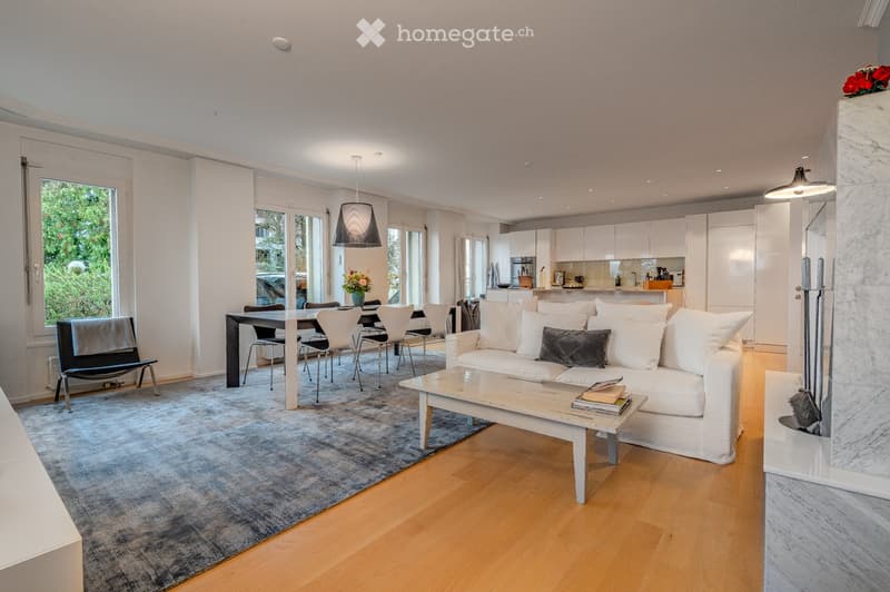 Modern open-plan living and dining area with light-colored furniture, a white sofa, coffee table, dining table with chairs, and a sleek kitchen area in the background.