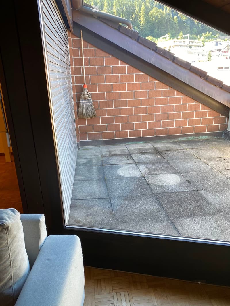 A modern living room with a grey sofa, wooden flooring, and a large window opening to a tiled roof terrace with brick walls and a broom leaning against it.