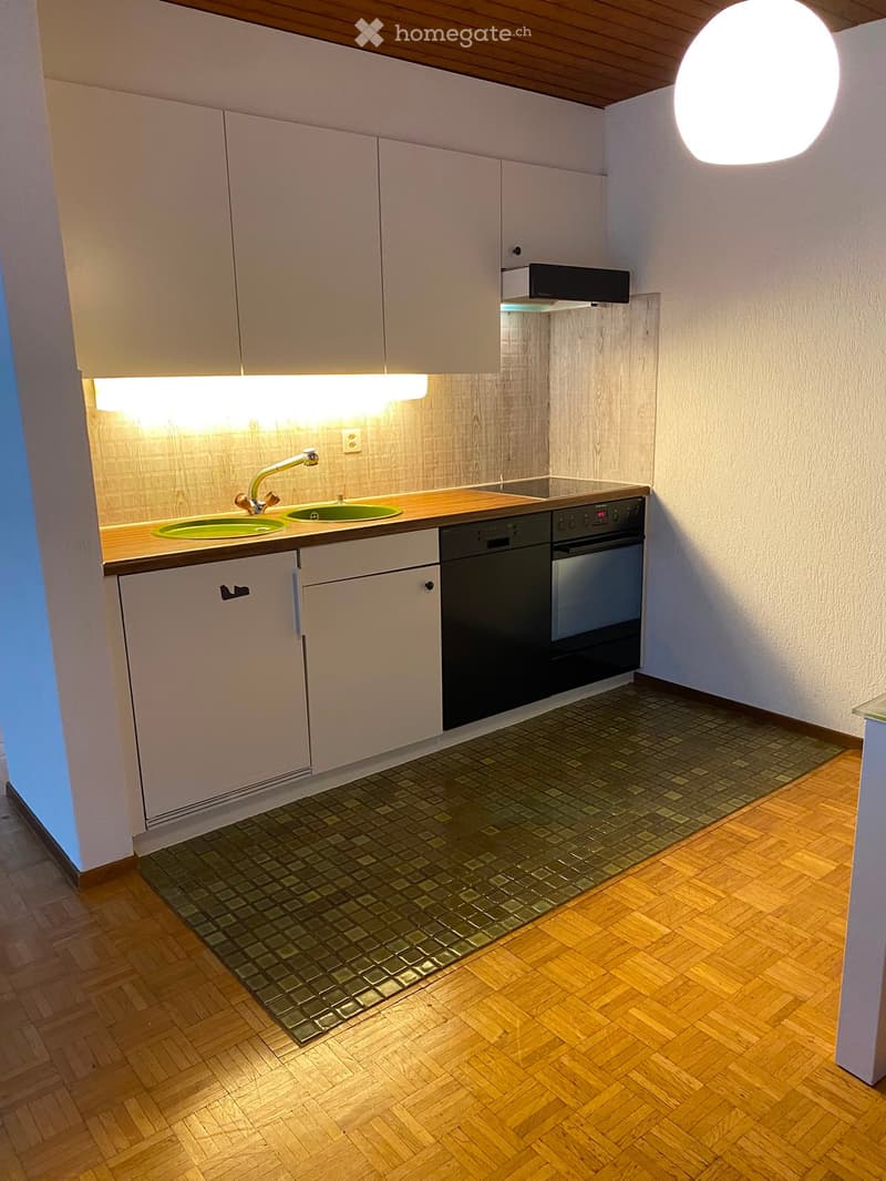 Modern kitchen with light-colored cabinets, a double sink, a dishwasher, and an oven. The floor has a mix of wooden and tiled sections.