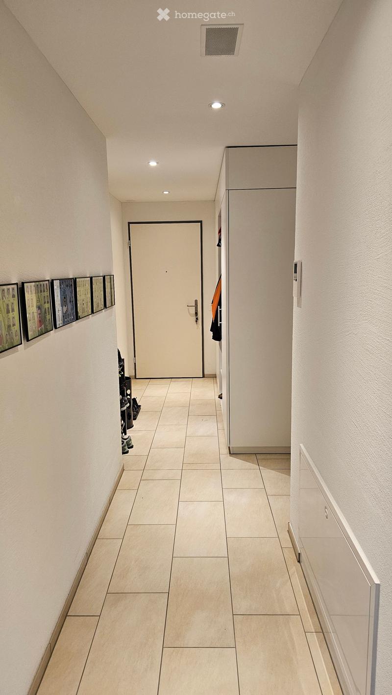 A clean, modern hallway with tiled flooring, white walls, recessed lighting, and a closed door at the end. A small shoe rack with shoes is visible on the left side, and framed pictures are mounted on the wall.