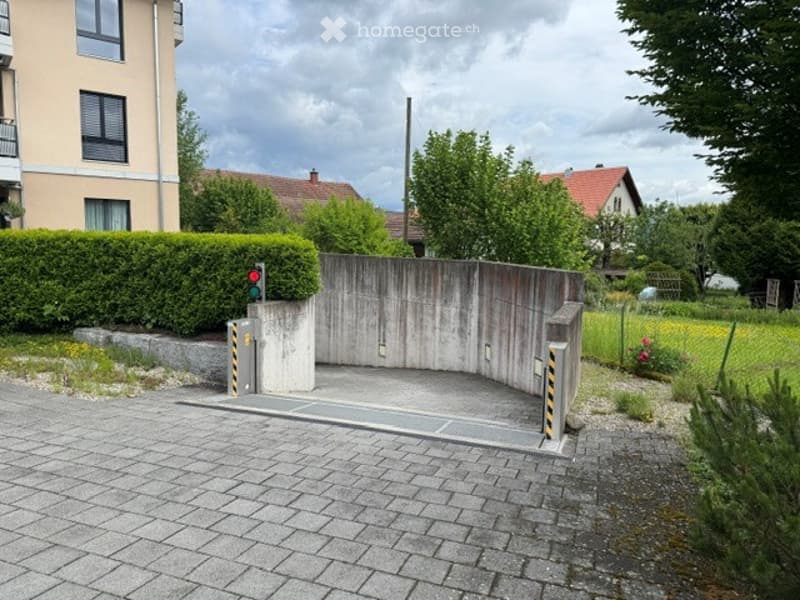 A gated entrance to a residential complex with a concrete barrier, traffic light, and surrounding greenery.