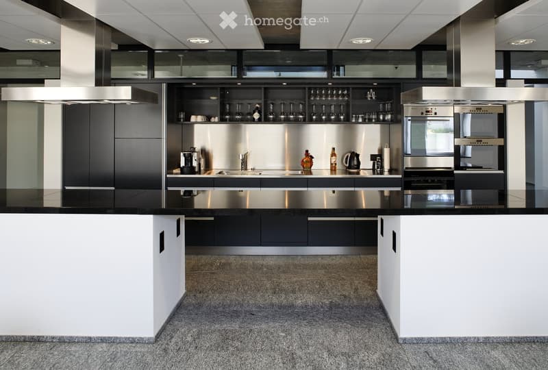 Modern kitchen with stainless steel appliances, black and white cabinetry, and a central island. Visible items include a coffee maker, oven, microwave, wine glasses, and a teapot.