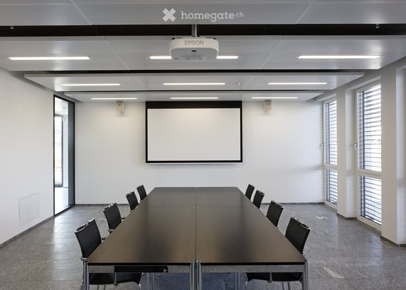 Modern conference room with a long wooden table, black chairs, a large screen, and a projector mounted on the ceiling.